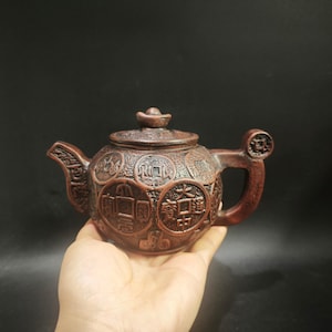 May include: A dark brown clay teapot featuring a textured surface decorated with relief carvings of circular ancient Chinese coins. The lid is topped with a small knob, and the handle incorporates a coin-shaped design element.