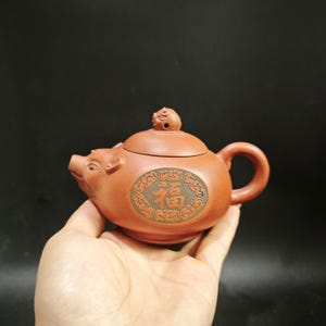 May include: A reddish-brown clay teapot shaped like a pig, featuring a small pig-shaped lid handle and a circular medallion on the side containing the Chinese character for good fortune, surrounded by a decorative border of swirling cloud patterns.