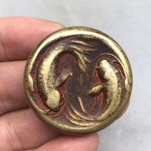 May include: A circular brass-toned metal button featuring a raised relief design of two koi fish swimming in a circular motion. The background of the relief is a deep reddish-brown color, while the fish figures are a metallic gold tone.