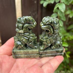 May include: Two dark green stone guardian lion figurines, each featuring intricate carved details, a wide open mouth, and a textured mane. The statues are positioned side by side, both mounted on square, flat bases with visible natural veining.