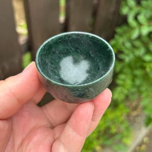 May include: A small, dark green jade bowl with a smooth, polished surface. The bowl is held in a hand, showcasing its rounded shape and the natural variations in the stone's colour. The bowl's interior has a lighter, almost white, pattern.