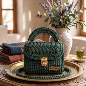 May include: A dark green crochet handbag featuring a thick braided top flap, a sturdy handle, and a gold-toned metal push-lock closure. A small rectangular wooden tag with the word Handmade is attached to the front of the bag.