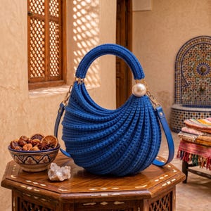 May include: A vibrant blue crescent-shaped handbag featuring a textured, ribbed crochet knit pattern. The bag includes a matching rounded handle, a large white pearl accent on one side, and a thin blue shoulder strap attached with gold-tone hardware.