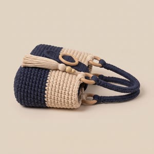 May include: A crochet handbag featuring alternating horizontal stripes of navy blue and cream yarn. The bag includes two dark blue crocheted handles attached with light-coloured wooden rings, a decorative wooden ring closure, and a cream-coloured tassel.
