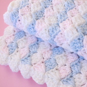 May include: A folded, handmade crochet blanket in a soft color palette of white, light blue, and pink. The blanket features a textured pattern, with each stitch creating a raised, shell-like design. The blanket is likely made of soft yarn.