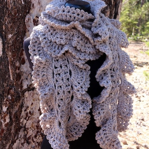Puede incluir: Una bufanda de ganchillo hecha a mano, de color gris claro, con un borde con volantes. La bufanda está drapeada sobre un maniquí negro, mostrando su intrincado diseño calado. La textura y el color sugieren un accesorio cálido y acogedor.