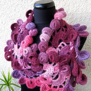 May include: A crocheted cowl in shades of pink and purple, with a textured, flower-like design. The cowl is displayed on a black mannequin.