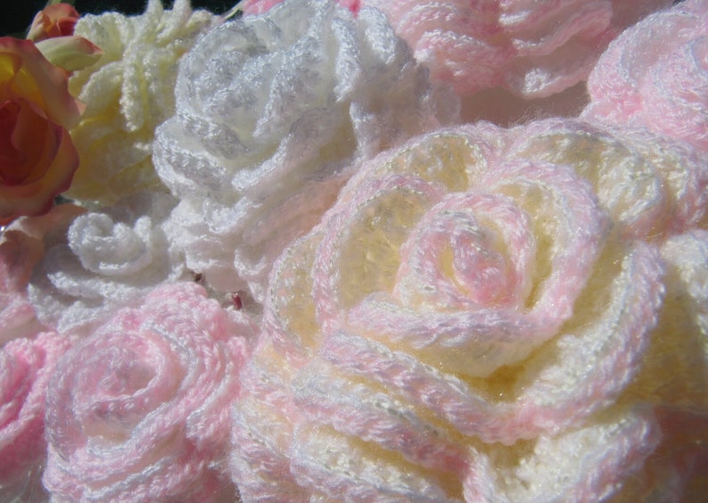 May include: A close-up of a bouquet of crocheted flowers in shades of white, pink, and cream. The flowers are made with a delicate, lacy stitch and have a soft, fluffy texture.