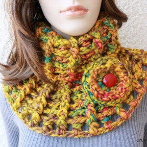 May include: A chunky, hand-crocheted cowl in shades of yellow, orange, and teal. The openwork design features a large, flower-shaped embellishment with a red button center. The cowl is a cozy accessory for cooler weather.