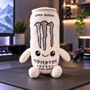 May include: A white plush toy shaped like a Monster Energy Ultra White drink can. It features the black Monster claw logo, the text ZERO SUGAR, MONSTER ENERGY, and ULTRA WHITE, along with a small embroidered smiling face with black eyes.