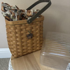 May include: A woven light brown basket with a dark green handle and a fabric liner featuring a pumpkin and leaf pattern. A small metal pumpkin charm is attached to the front. A clear plastic rectangular cover sits beside the basket.