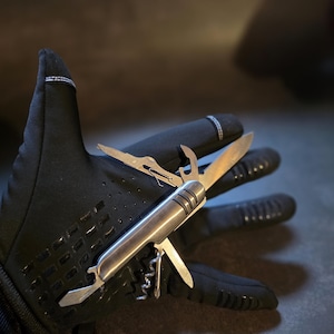 Op de afbeelding: Een zilverkleurig multifunctioneel zakmes met diverse uitgeklapte messen en gereedschappen, waaronder een kurkentrekker en een flesopener, rustend op een zwarte sporthandschoen met textuur voor extra grip op de handpalm en vingers.