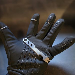 May include: A black glove with a textured grip pattern on the palm and fingers, featuring a silver-coloured metal multi-tool resting across the palm. The tool has a flat, notched edge, a series of diagonal cutouts, and a hexagonal opening at one end.