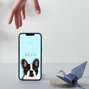 May include: A smartphone with a light blue screen displaying the name LUCA and a black and white French Bulldog illustration. Next to the phone is a blue paper crane on a small circular gray base. The phone shows the time 11:47 on the display.