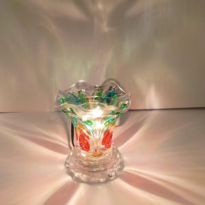 Puede incluir: Un calentador de cera eléctrico de vidrio transparente con un borde superior ondulado y detalles en verde y rojo. La base es circular y transparente, y la fuente de luz interna proyecta patrones de luz radiantes en el área circundante.