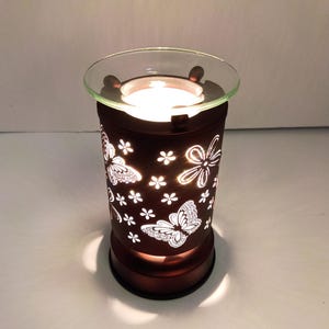 Puede incluir: Un calentador de cera eléctrico cilíndrico de color marrón oscuro con diseños recortados de mariposas y flores pequeñas. El dispositivo tiene un plato de vidrio transparente en la parte superior y emite una luz cálida a través de los patrones.
