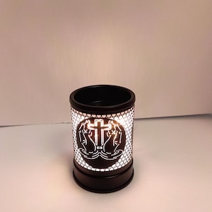May include: A cylindrical black wax warmer featuring a cut-out design of praying hands and a cross. The metal exterior has a lattice pattern that allows light to shine through from the interior, creating a glowing effect against the dark surface.
