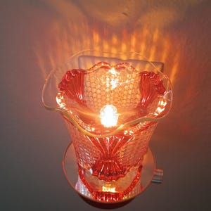 May include: A plug-in wall warmer featuring a red glass dish with a textured honeycomb pattern and a ruffled, scalloped rim. The glass is illuminated from within by a bright light, casting a warm glow and intricate light patterns onto the wall.