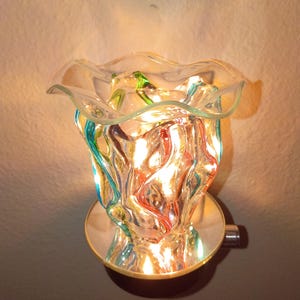 May include: A plug-in wax warmer featuring a clear, ruffled glass bowl with swirling red, blue, and green accents. The glass vessel sits atop a circular, reflective base that illuminates from within, casting a warm glow through the colorful glass.