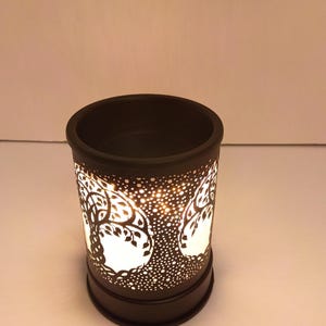 May include: A cylindrical black wax warmer featuring a cut-out design of a tree with intricate branches and leaves. The exterior has a perforated pattern that allows light to shine through from the interior, creating a glowing effect.
