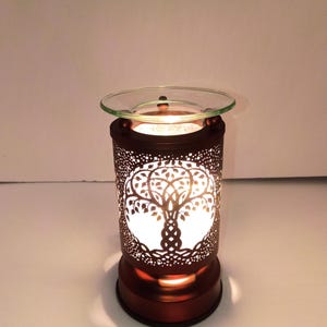 May include: A cylindrical metal wax warmer featuring a dark brown finish and intricate laser-cut tree of life patterns. The device includes a clear glass dish on top and emits a warm, bright glow from the internal light source through the cutouts.