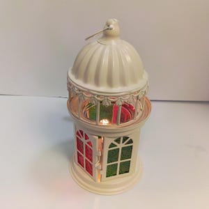 May include: A cream-colored metal lantern with a domed top and a small hanging loop. The lantern features a hinged door and side panels with translucent red and green patterned glass inserts, allowing light to shine through the structure.