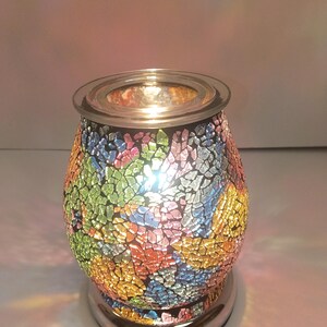 May include: An electric wax warmer featuring a rounded, bulbous body covered in a multi-colored mosaic of glass shards in shades of green, blue, pink, and yellow. The top includes a clear glass dish, and the base is a polished silver metal.