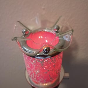 May include: A plug-in wax warmer featuring a cylindrical base with a pink crackle glass finish and a silver-toned metal rim. The top consists of a clear, ruffled glass dish designed to hold wax, illuminated from within by a bright pink light.