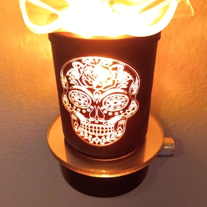 Könnte beinhalten: Ein schwarzer elektrischer Wachswärmer mit einem leuchtenden weißen Totenkopf-Design auf der Vorderseite. Der zylindrische Wärmer hat eine gewellte, durchscheinende Schale, die ein warmes, helles orangefarbenes Licht ausstrahlt und das florale Muster beleuchtet.