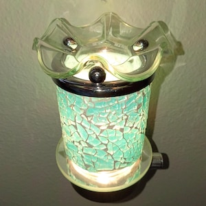 May include: A plug-in wax warmer featuring a cylindrical body covered in turquoise mosaic glass tiles. The top has a clear, ruffled glass dish held by a silver-toned metal rim. The unit emits a warm glow from within, illuminating the glass.