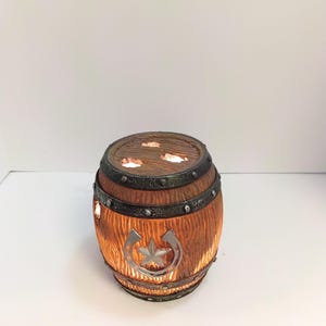 May include: A brown barrel-shaped lamp with a textured wood grain finish, featuring dark metal-colored bands with rivets. A silver-colored horseshoe and star emblem are centered on the front. Light glows from within the barrel and through holes in the top.