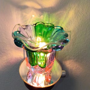 May include: A plug-in wall lamp featuring a ruffled, flower-shaped glass shade in vibrant shades of green, purple, and clear glass. The base is circular and reflective, casting a warm, illuminated glow that projects colorful light patterns.