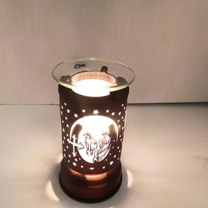 Puede incluir: Un calentador de cera cilíndrico de color marrón oscuro con un diseño recortado de una cabeza de caballo, una cruz y numerosas estrellas pequeñas. El dispositivo emite una luz blanca brillante y tiene un plato de vidrio transparente encima.