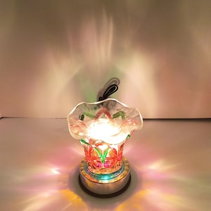 May include: A glass wax warmer featuring a ruffled top edge and a clear body with red and green decorative accents. The base is circular and metallic, emitting a bright light that casts colorful, prismatic reflections onto the surrounding area.