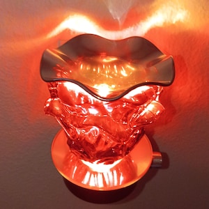 May include: A plug-in wax warmer featuring a vibrant red glass body with a textured, organic shape and a smooth, ruffled top dish. The device emits a warm, bright red glow from an internal light source, casting a soft illumination outward.