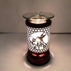 May include: A cylindrical electric wax warmer with a dark metallic finish featuring a cutout pattern of a hummingbird hovering near flowers. The device emits a warm white glow from within and includes a clear glass dish on top for wax melts.