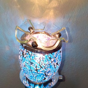 May include: An electric wax warmer featuring a blue mosaic glass body with a crackled texture. The top consists of a clear, ruffled glass dish held by a silver-toned metal rim. The device emits a bright internal glow, illuminating the blue glass.