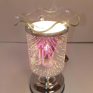 Puede incluir: Un calentador de cera eléctrico cilíndrico con base cromada y cuerpo de cristal espejado que crea un efecto de luz infinita. La parte superior tiene un plato de cristal transparente con bordes ondulados para cera, emitiendo luz.