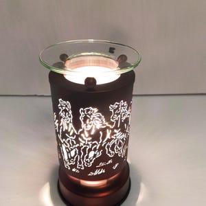 May include: A cylindrical dark brown wax warmer featuring a cut-out design of running horses. The metal body allows light to shine through the horse silhouettes, and it is topped with a clear glass dish for holding wax melts, illuminated from within.
