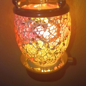 May include: A plug-in wax warmer featuring a cylindrical body covered in a mosaic of orange, yellow, and red glass shards. The top section has a clear, ruffled glass dish held by a dark metal frame, illuminated from within by a warm light.