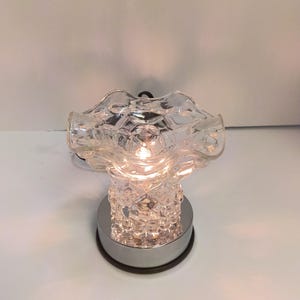 May include: A clear glass wax warmer featuring a ruffled top bowl and a textured, diamond-patterned base. The unit sits on a circular, reflective chrome-finished pedestal and contains an illuminated light bulb visible through the glass structure.