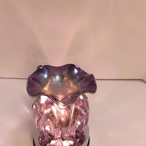 May include: A purple glass wax warmer featuring a ruffled top edge and a textured, bulbous body. The piece sits on a circular silver-toned base and emits a warm glow from within, highlighting the iridescent finish of the glass material.