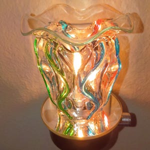 May include: A plug-in glass wax warmer featuring a ruffled top edge and a clear body with swirling vertical ribbons of green, blue, and orange glass. The interior is illuminated by a warm light, casting a glow through the textured glass walls.