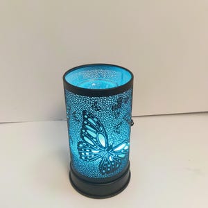 Puede incluir: Un calentador de cera eléctrico cilíndrico con un exterior de metal negro que presenta un patrón de mariposas y mosaico cortado con láser. El interior brilla con una luz azul intensa, iluminando los recortes sobre el marco oscuro.