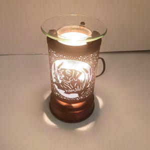 Könnte beinhalten: Ein zylindrischer Wachswärmer aus Metall mit bronzefarbener Oberfläche und einer ausgeschnittenen Hundesilhouette. Der Wärmer hat eine klare Glasschale auf der Oberseite und strahlt durch die Muster ein warmes Licht aus.