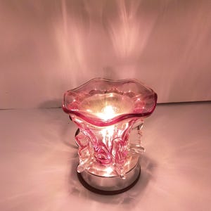 Puede incluir: Un calentador de cera de vidrio rosa con una parte superior ondulada en forma de cuenco y un cuerpo texturizado y escultural. El dispositivo está iluminado desde el interior, proyectando un brillo cálido y patrones de luz suave desde su base.