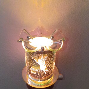May include: A plug-in wax warmer featuring a cylindrical glass body with a reflective, multi-faceted interior surface. A white silhouette of a fairy and the text "Make a wish" are visible on the front. The top has a clear, ruffled glass dish.