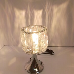 May include: A table lamp with a cylindrical, textured clear glass shade and a polished chrome base. The lamp is illuminated, casting light patterns onto the wall behind it. A black power cord extends from the base of the lamp.