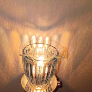 May include: A plug-in wax warmer featuring a clear, ribbed glass bowl with a ruffled top edge. The device is illuminated from within, casting a warm, golden glow and light patterns onto the wall behind it, with a circular base attached to the wall.