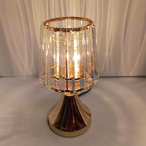 Może przedstawiać: Lampa stołowa z podstawą z polerowanego metalu w kolorze złotym oraz cylindrycznym kloszem wykonanym z pionowych, przezroczystych, kryształowych prętów szklanych. Lampa emituje ciepłe światło, rzucając miękkie, liniowe cienie.